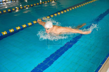Téléchargez les photos : Sportif adulte en casquette blanche engagé dans des exercices dans l'eau, nageant dans le style de papillon - en image libre de droit