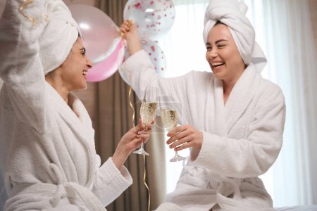 Porträt glücklicher Frauen im Bademantel im luxuriösen Hotelzimmer und klirrenden Gläsern mit Sekt