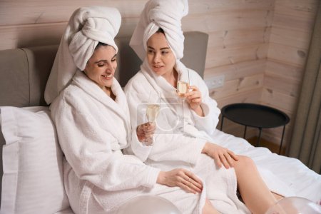 Téléchargez les photos : Deux dames portant des peignoirs allongés dans un grand lit dans la chambre d'hôtel, parlant et tenant des lunettes avec du vin mousseux - en image libre de droit