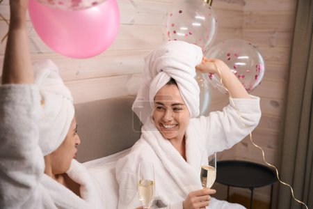 Téléchargez les photos : Deux femmes heureuses en robes blanches assises sur le lit avec des ballons et des verres de champagne et riant pendant la fête de poule - en image libre de droit