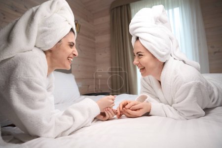 Téléchargez les photos : Deux jolies jeunes femmes ayant une journée de fille, portant des peignoirs blancs et des serviettes, allongées sur le lit tout en riant - en image libre de droit