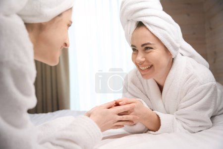 Téléchargez les photos : Deux jolies jeunes femmes ayant une journée de fille, portant des peignoirs blancs et des serviettes, s'allongeant sur le lit et se tenant la main - en image libre de droit