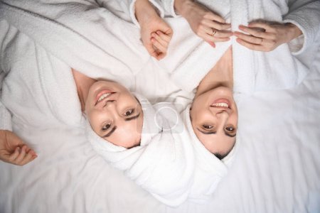 Téléchargez les photos : Portrait de deux femmes en peignoirs blancs allongées au lit satisfaites des soins du corps et regardant la caméra - en image libre de droit