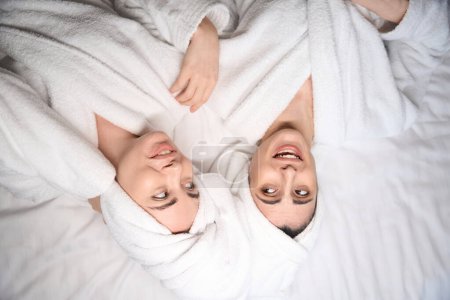 Téléchargez les photos : Portrait de deux femmes en peignoirs blancs couchées au lit satisfaites des soins corporels - en image libre de droit