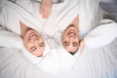 Téléchargez les photos : Vue du dessus de deux dames en peignoirs blancs allongées au lit satisfaites des soins du corps et riant - en image libre de droit