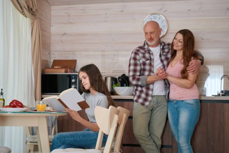 Téléchargez les photos : Adolescente concentrée assise à la table de cuisine faisant ses devoirs sous la supervision parentale - en image libre de droit