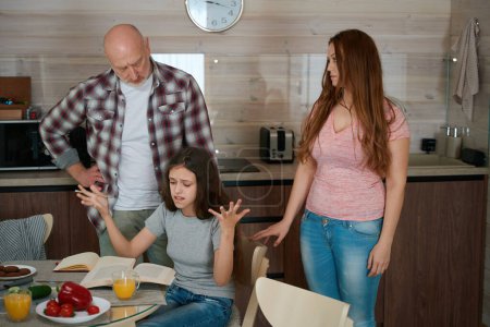 Photo for Homeschooler seated at kitchen table throwing her hands in air while looking task in textbook in presence of serious parents - Royalty Free Image