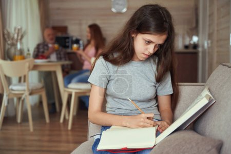 Téléchargez les photos : Adolescente concentrée assise sur le canapé faisant ses devoirs pendant que maman et père buvaient du jus d'orange à table - en image libre de droit