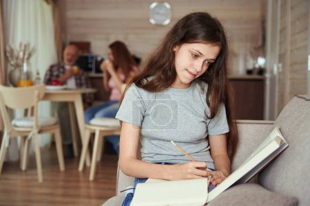 Téléchargez les photos : Calme concentré éducateur assis sur le canapé faire ses devoirs tandis que les parents buvant du jus d'orange à la table de cuisine - en image libre de droit
