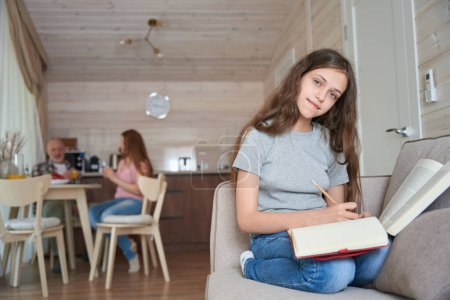 Téléchargez les photos : Une adolescente souriante assise sur un canapé faisant ses devoirs pendant que papa et maman buvaient du jus d'orange dans la cuisine - en image libre de droit