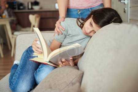 Téléchargez les photos : Jeune femme concentrée allongée sur le canapé tout en lisant le livre en présence de la mère - en image libre de droit