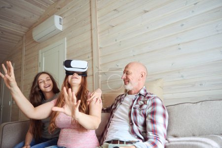 Foto de Mujer con auriculares VR jugando videojuegos mientras está sentada en el sofá en compañía de su complacido esposo y su hija - Imagen libre de derechos