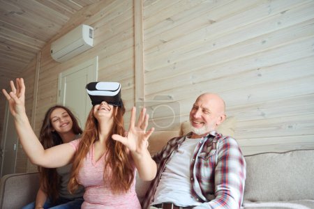 Téléchargez les photos : Dame agréable dans VR lunettes de jeu vidéo tout en étant assis sur le canapé en présence de joyeux conjoint et fille - en image libre de droit