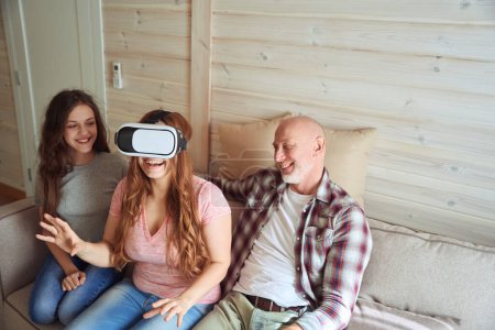 Foto de Alegre ama de casa en auriculares VR jugando videojuegos mientras está sentada en el sofá con su hija sonriente y su marido - Imagen libre de derechos