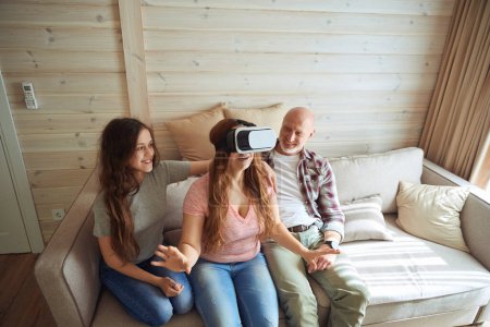 Téléchargez les photos : Joueuse en lunettes VR assise sur un canapé jouant à un jeu vidéo entouré d'une fille et d'un conjoint joyeux - en image libre de droit