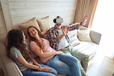 Téléchargez les photos : Joueur agréable dans VR lunettes de jeu vidéo tout en étant assis sur le canapé avec sa femme heureuse et sa fille - en image libre de droit
