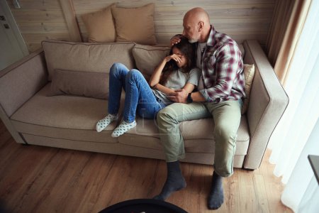 Photo for Loving father seated on sofa in living room hugging his depressed adolescent kid - Royalty Free Image