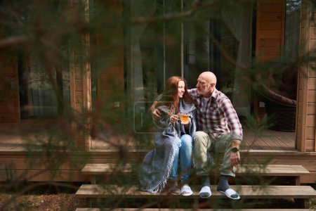 Foto de Amante esposo abrazando sonriente esposa con taza de té en las manos sentadas a su lado en escalones de madera del porche - Imagen libre de derechos