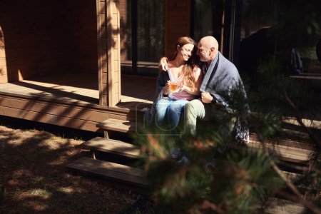 Foto de Hombre abrazando a su alegre esposa sentada junto a él en el porche con una taza de bebida herbal en las manos - Imagen libre de derechos