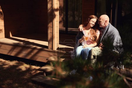 Foto de Romántico macho abrazando a su feliz esposa sentada junto a él en el porche con una copa de bebida herbal en las manos - Imagen libre de derechos