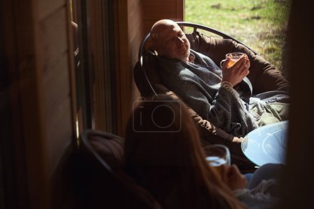 Téléchargez les photos : Agréable mâle et sa compagne assis dans des chaises tout en parlant sur la véranda avec des tasses de boisson à base de plantes dans les mains - en image libre de droit