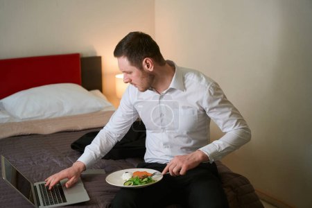 Schwerwiegender junger Mann sitzt auf Bett im Hotelzimmer, sticht mit Gabel Essen und tippt auf Laptop-Tastatur