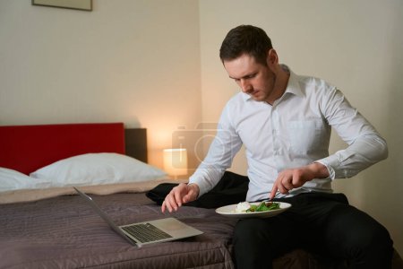 Male pricking food with fork while sitting on bed in front of portable computer in aparthotel