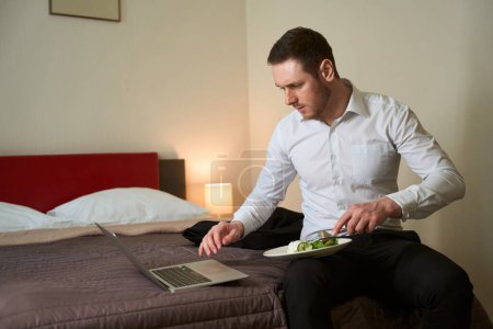 Foto de Hombre de negocios sentado en la cama en la habitación del hotel pinchando comida con tenedor mientras mira la pantalla del ordenador portátil - Imagen libre de derechos