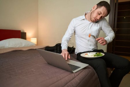 Foto de Joven emprendedor sentado en la cama en aparthotel mientras escribe en el teclado de la computadora durante la llamada telefónica - Imagen libre de derechos