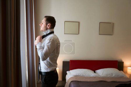 Foto de Vista lateral de un joven elegante atando corbata alrededor del cuello de la camisa mientras mira a través de cortinas de ventana en suite de lujo - Imagen libre de derechos