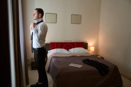 Photo for Side view of pensive businessman tying his necktie around shirt collar while looking through hotel window curtains - Royalty Free Image