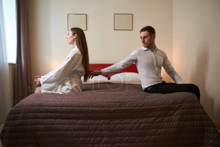 Sad young man seated on bed in luxury suite touching hair of his offended female companion