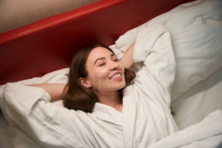 Téléchargez les photos : Souriant heureux femelle en peignoir couché avec les yeux fermés dans un lit confortable - en image libre de droit