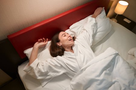 Photo for Pleased woman wrapped in blanket lying on soft white pillows in bed in suite - Royalty Free Image