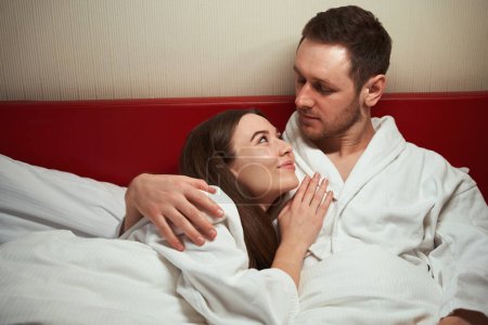 Photo for Romantic guy hugging young woman lying on his shoulder in bed in hotel room - Royalty Free Image