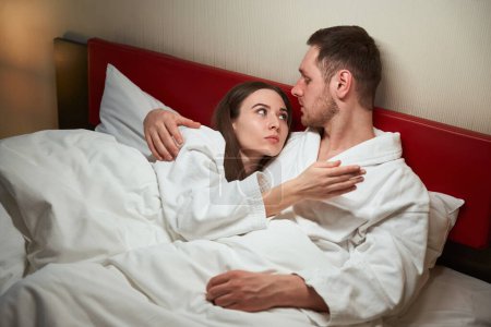 Foto de Hombre abrazando a su compañera seria acostado en su hombro en la cama en la habitación de hotel durante la conversación - Imagen libre de derechos