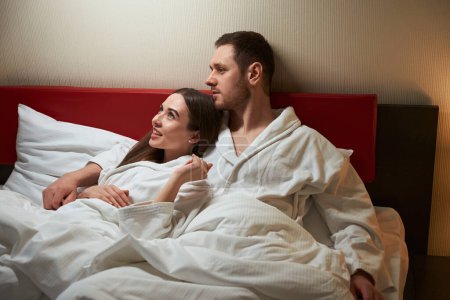 Téléchargez les photos : Souriant heureux jeune femme couchée dans le lit dans les bras de son cher compagnon en suite - en image libre de droit