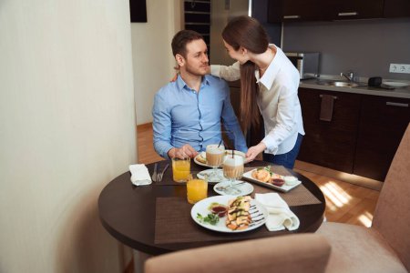 Téléchargez les photos : Homme assis à table avec de la nourriture et regardant la femme dans la chambre du motel - en image libre de droit