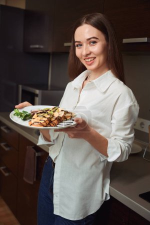 Photo for Happy female standing near table and holding plate of food in hotel room, looking at the camera - Royalty Free Image