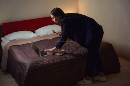 Téléchargez les photos : Jeune femme en costume d'affaires debout près du lit et dîne, tapant sur ordinateur portable dans la chambre d'hôtel - en image libre de droit