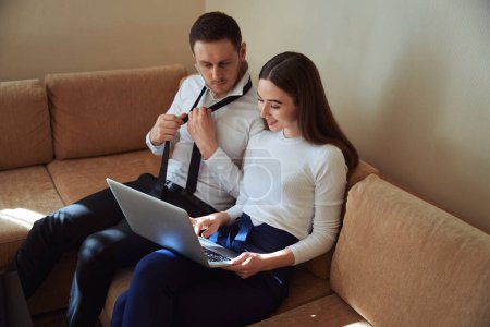 Téléchargez les photos : Homme assis sur un canapé près d'une femme travaillant sur un ordinateur portable et s'habillant dans le motel - en image libre de droit
