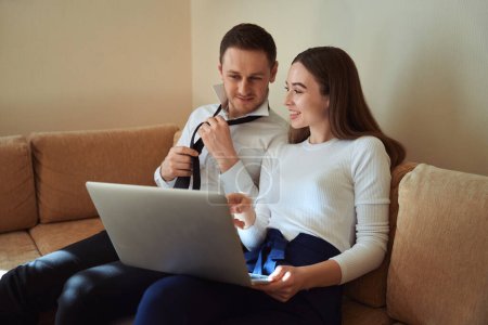 Photo for Man sitting on sofa near smiling female and tying his tie, looking at laptop in the hotel - Royalty Free Image