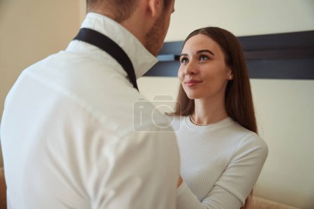 Photo for Smiling female standing near young man and tying tie in the motel room - Royalty Free Image