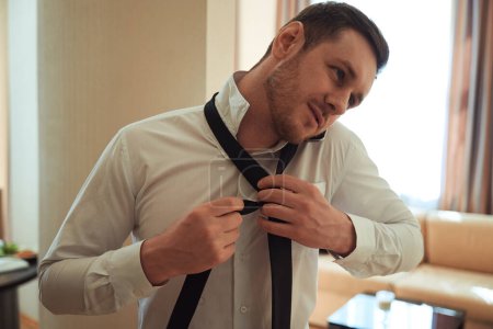 Téléchargez les photos : Jeune homme appelant sur smartphone et attachant sa cravate dans la chambre du motel - en image libre de droit