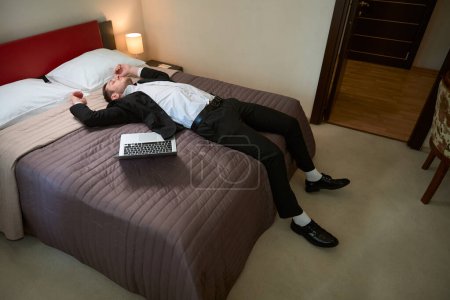 Téléchargez les photos : Jeune homme en costume d'affaires reposant sur le lit près d'un ordinateur portable dans la chambre d'hôtel - en image libre de droit