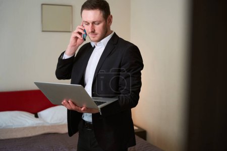 Photo for Male in business suit with computer in his hand calling on phone in the hotel room - Royalty Free Image