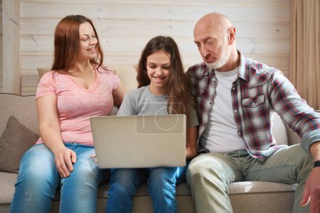 Téléchargez les photos : Joyeuse fille assise sur le canapé faisant des devoirs sur ordinateur portable en présence de sa mère souriante et perplexe père - en image libre de droit