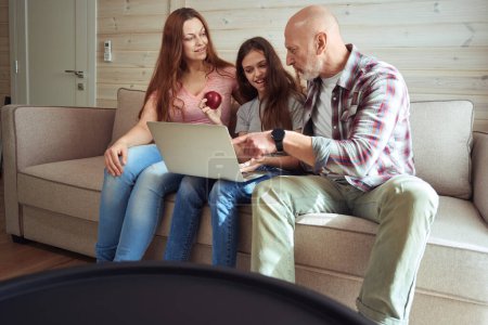 Photo for Serious man seated on sofa pointing at laptop screen to his smiling daughter and wife - Royalty Free Image