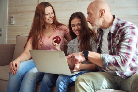 Photo for Focused man seated on sofa showing something on computer monitor to his joyous daughter and spouse - Royalty Free Image