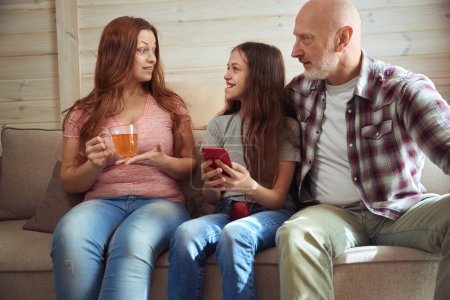 Photo for Woman with teacup sitting on sofa beside her cheerful daughter with smartphone and calm husband - Royalty Free Image